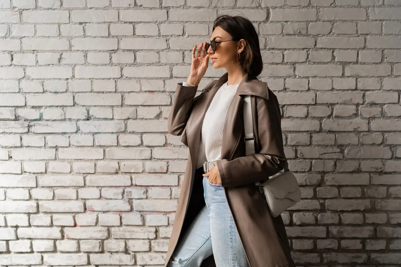 Modische, kurzhaarige Frau in lässigem Ledermantel und Jeans posiert vor einer städtischen Backsteinwand.